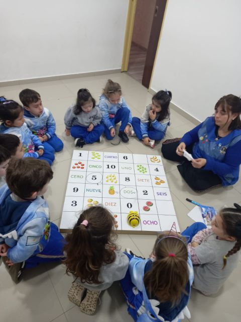 La docente y los chicos, en ronda alrededor de un tablero con números y frutas, con un robot de piso